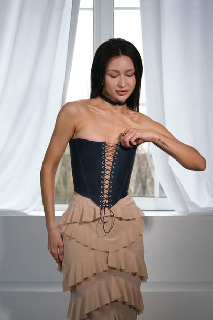 A woman wearing a black corset and a beige ruffled skirt stands in front of a window, looking down at her hand.