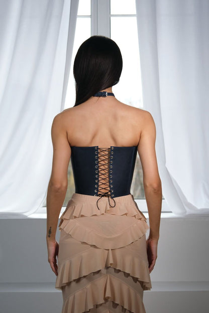 A woman wearing a black corset and a beige ruffled skirt stands in front of a window, facing away from the camera.