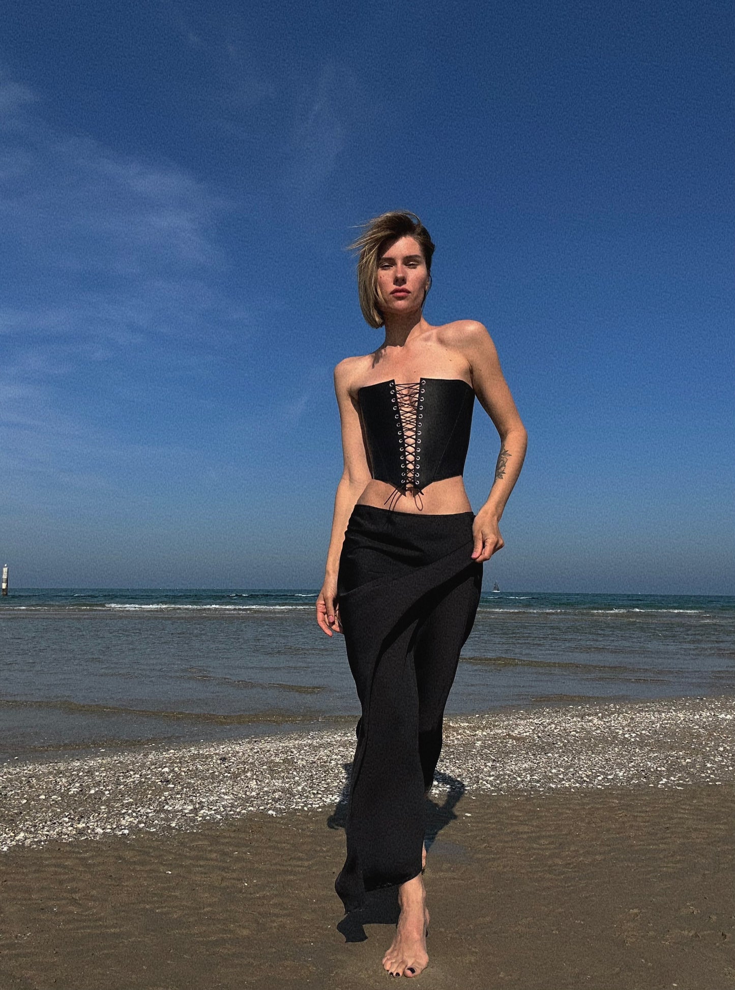 A woman in a black strapless top and skirt walks along a beach, with the ocean and sky visible in the background.