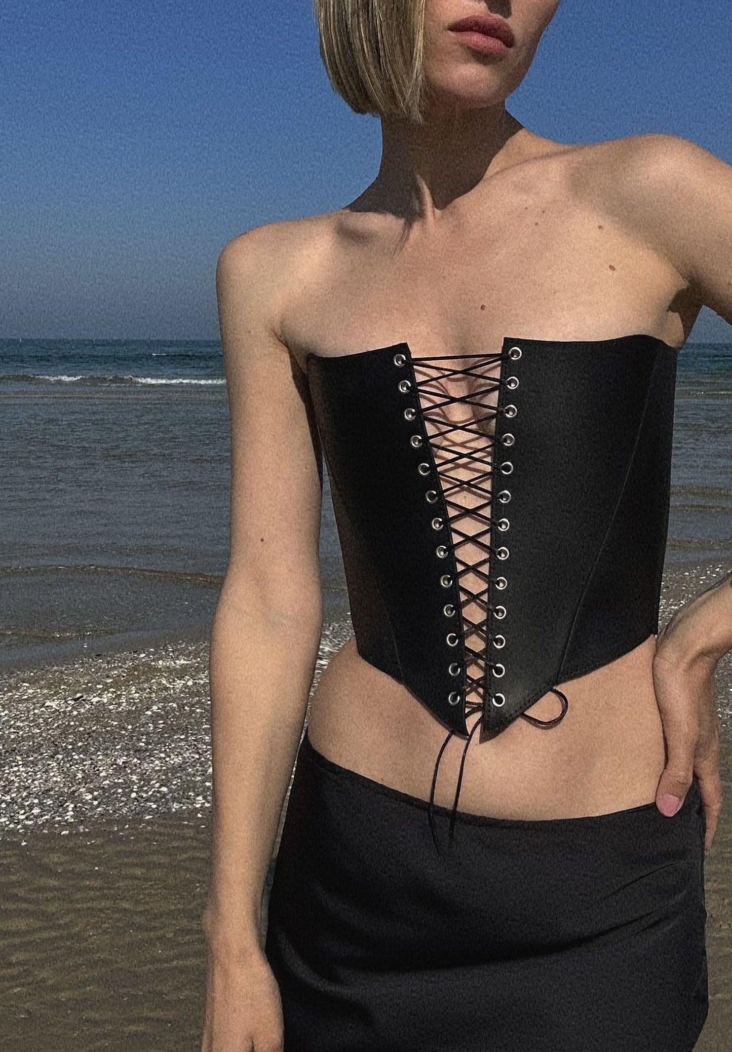 A woman wearing a strapless black corset stands on a beach, with the ocean visible in the background.
