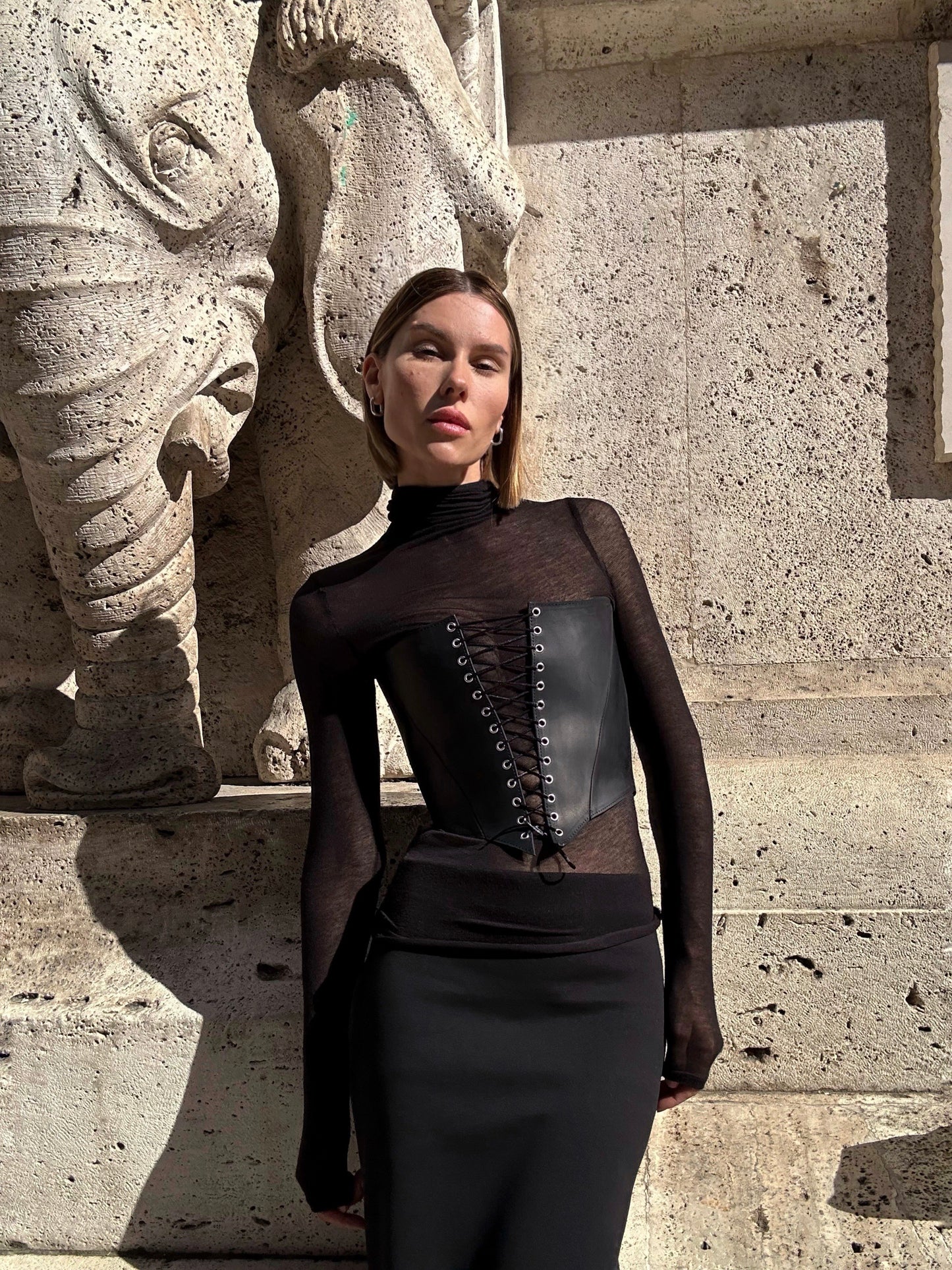 A woman in a black outfit stands in front of a stone wall, with a statue visible behind her.