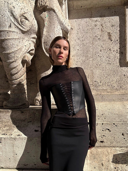 A woman in a black outfit stands in front of a stone wall, with a statue visible behind her.