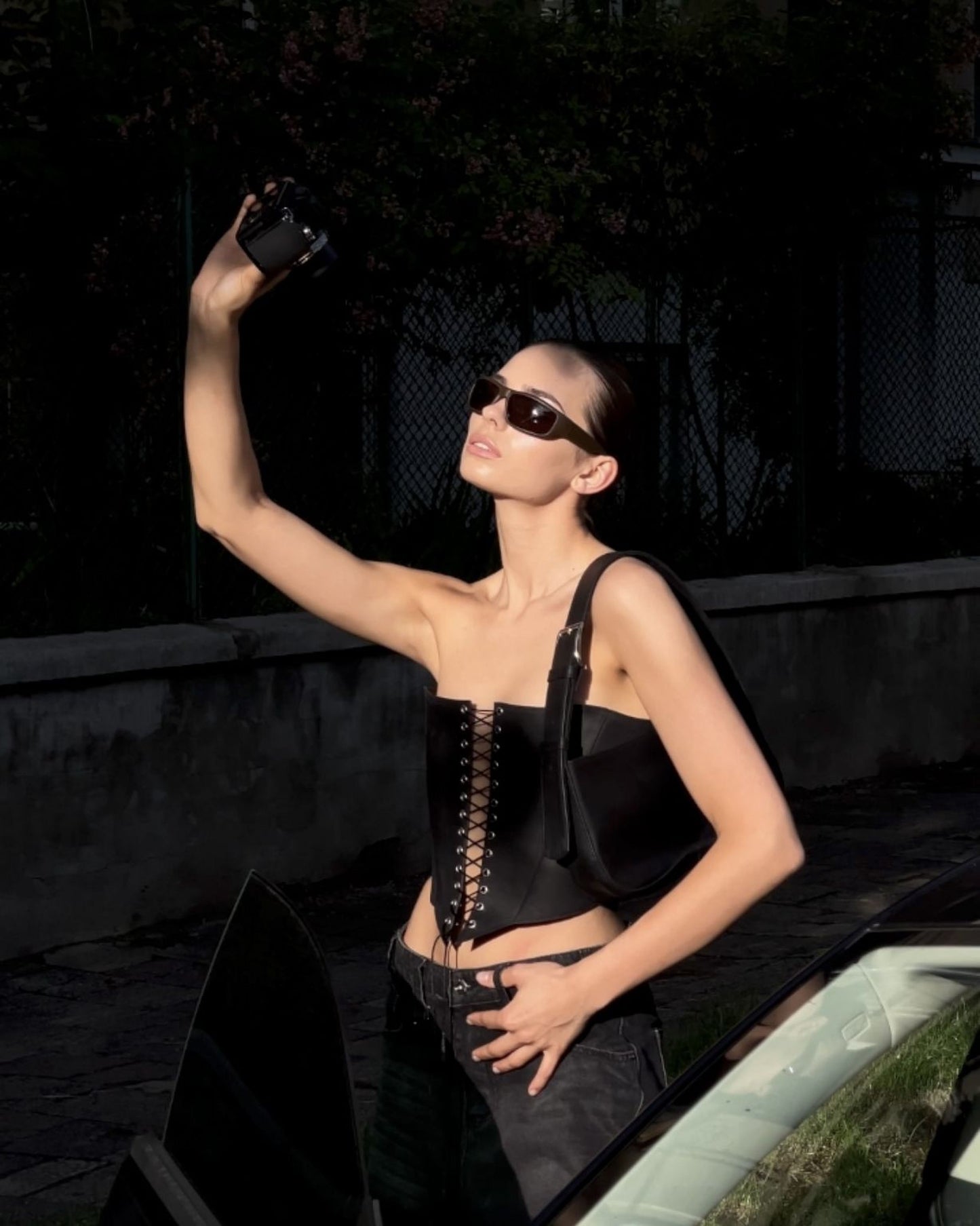 A woman wearing sunglasses and a black outfit is taking a selfie with her arm raised, holding a camera in her hand.