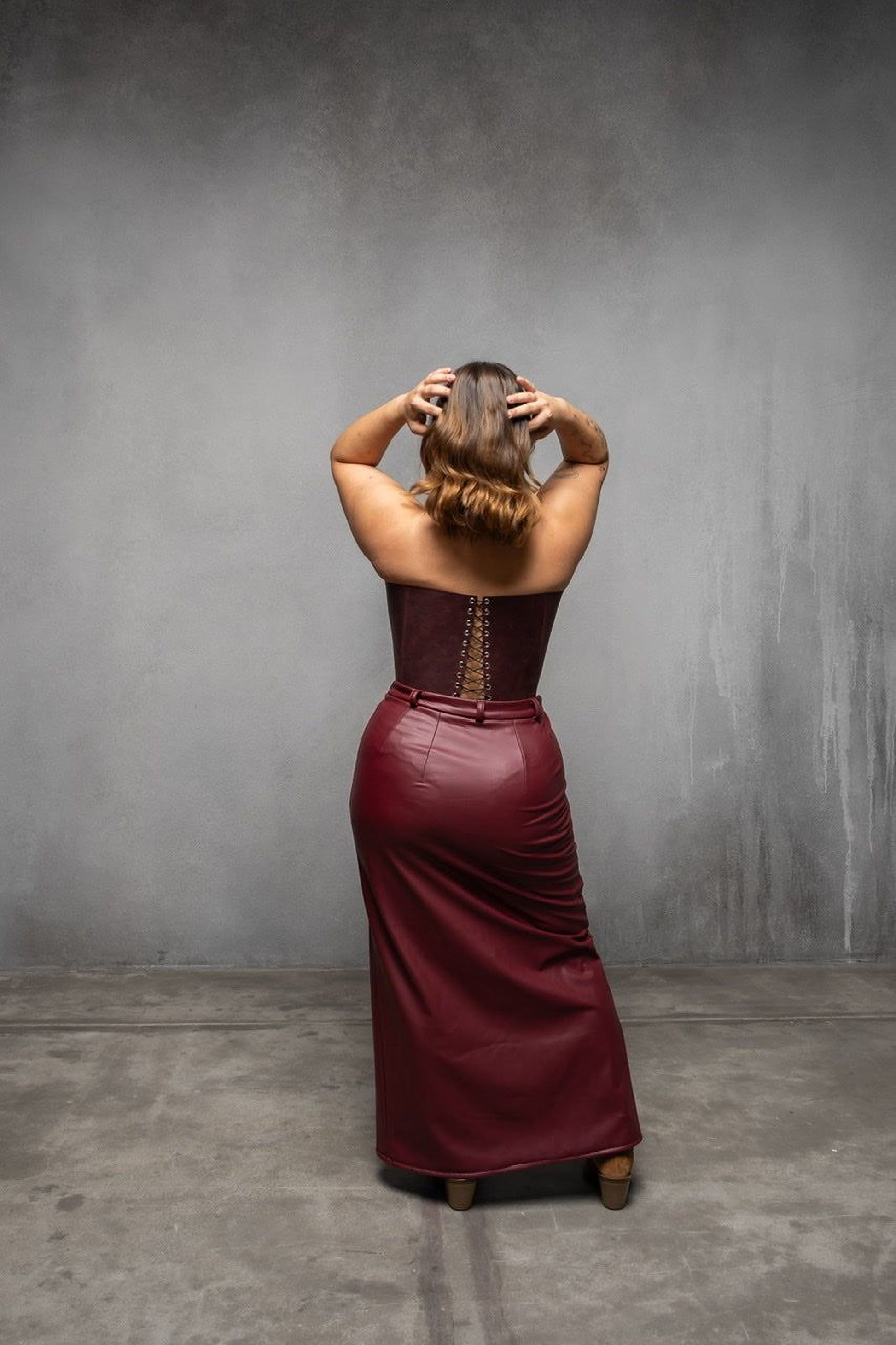 A woman in a burgundy dress stands with her hands on her head, facing away from the camera, against a gray background.