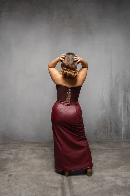 A woman in a burgundy dress stands with her hands on her head, facing away from the camera, against a gray background.
