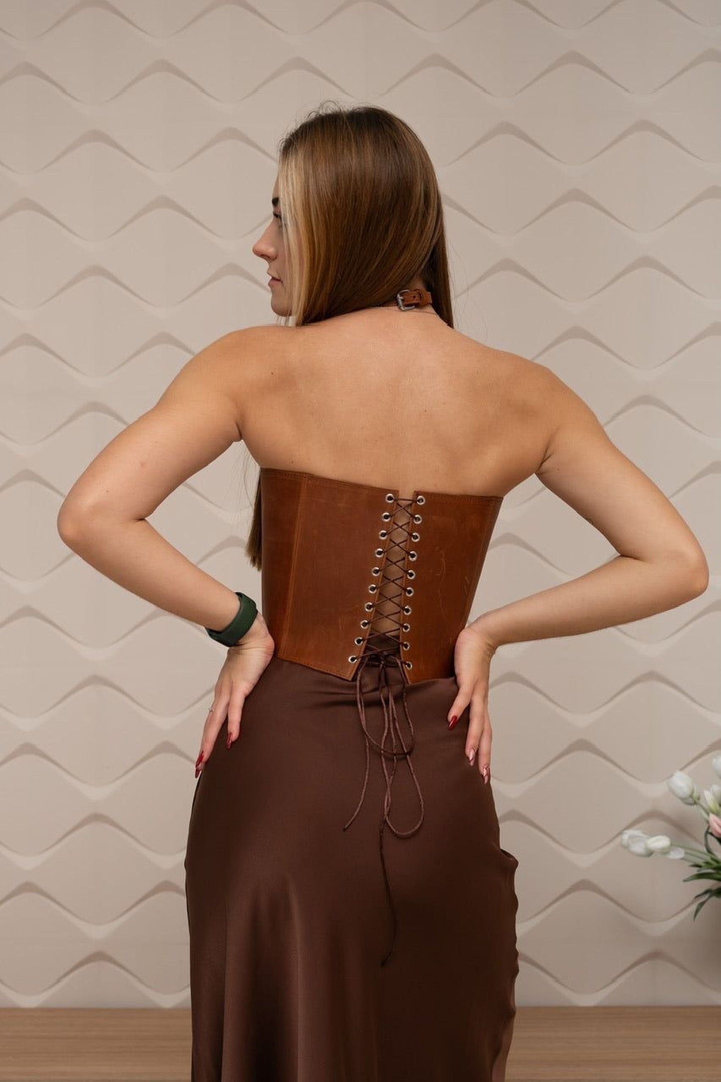 A woman wearing a brown leather corset stands with her hands on her hips, facing away from the camera.