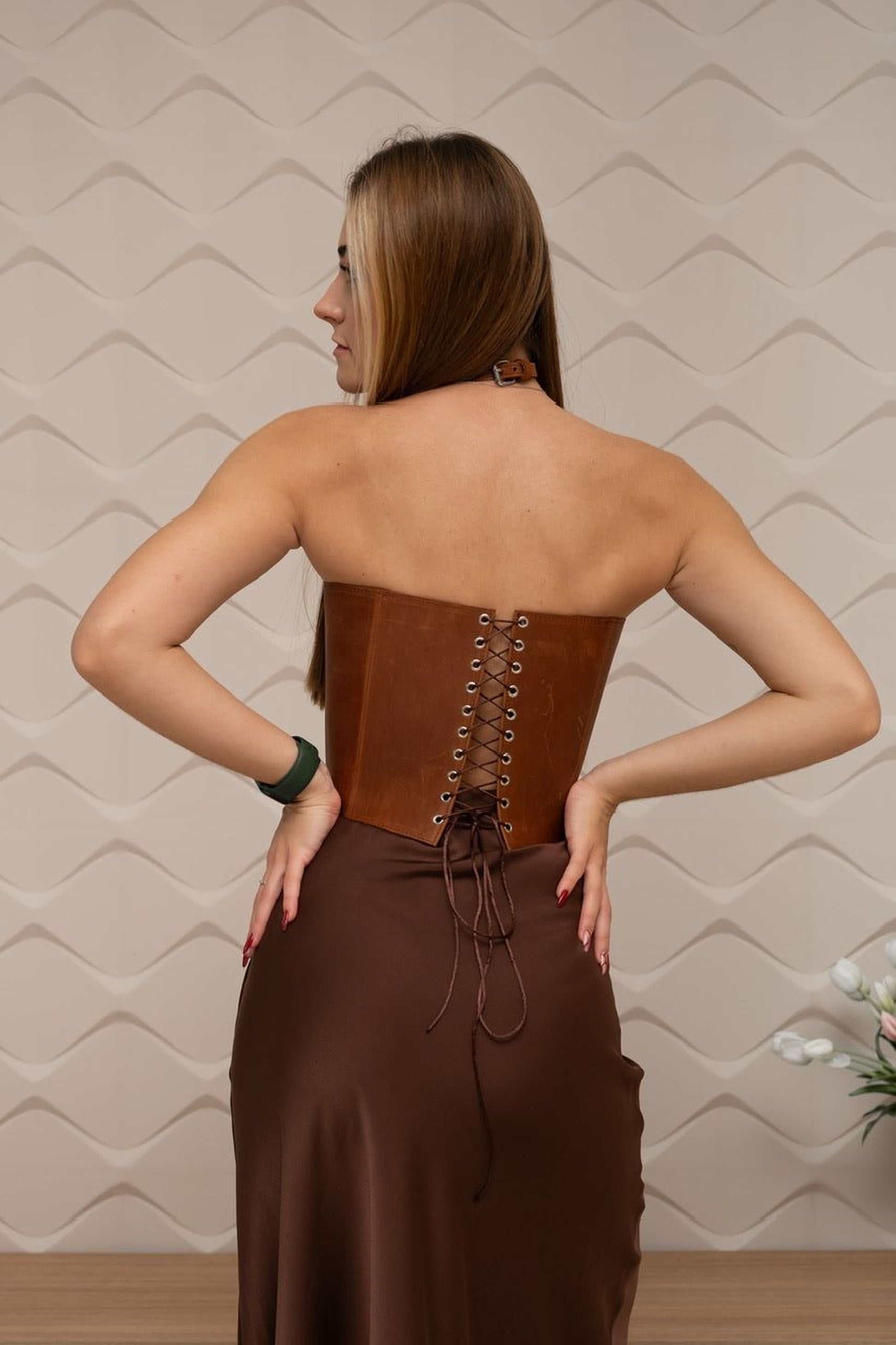 A woman wearing a brown leather corset stands with her hands on her hips, facing away from the camera.